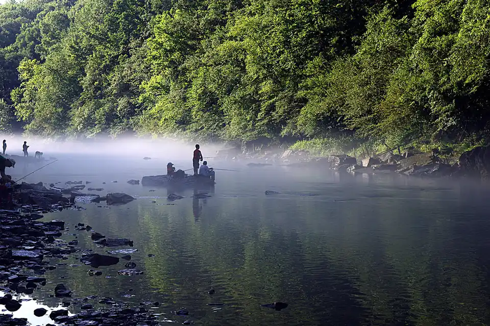 Several anglers fish from the shore and a small boat on a misty, tree-lined river.