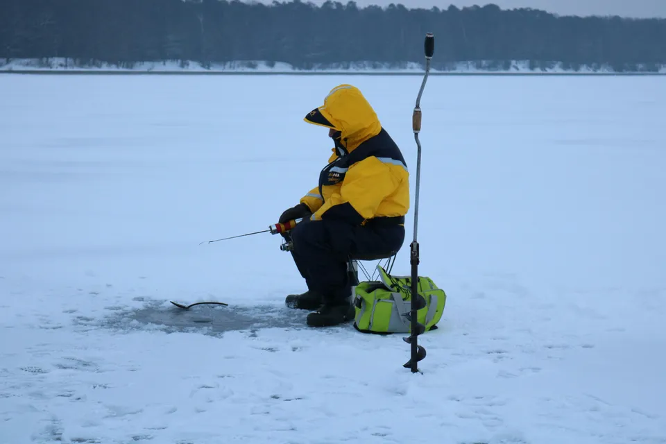An angler on a stool uses a short rod by a hole, with a hand ice auger standing beside him.