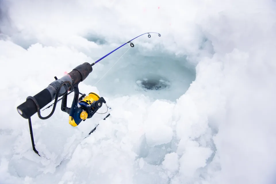 A short ice fishing rod with a small spinning reel resting on its stand beside a freshly drilled hole in snowy ice.