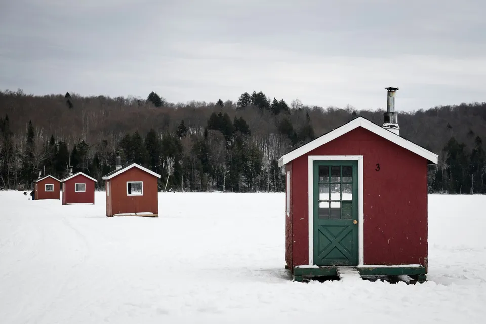 A row of red wooden ice fishing shanties with stovepipes on a snow-covered lake.