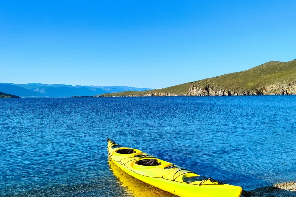 A yellow tandem kayak rests at the shore of a wide, clear blue lake with a rocky headland and distant mountains under a clear sky.