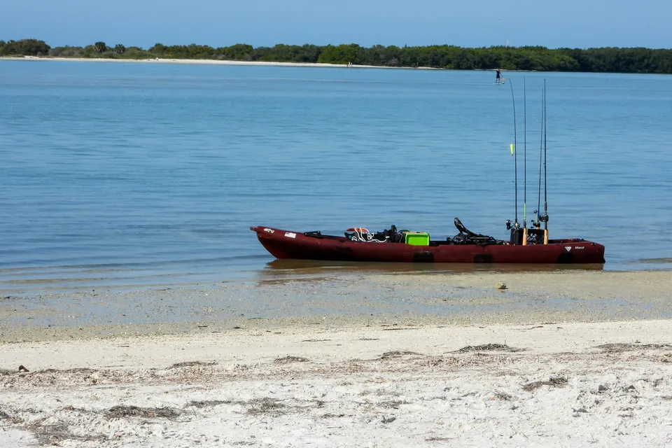 A dark red fishing kayak with a frame seat, a crate and four rods in holders sits at the edge of calm, shallow water on a sand beach.