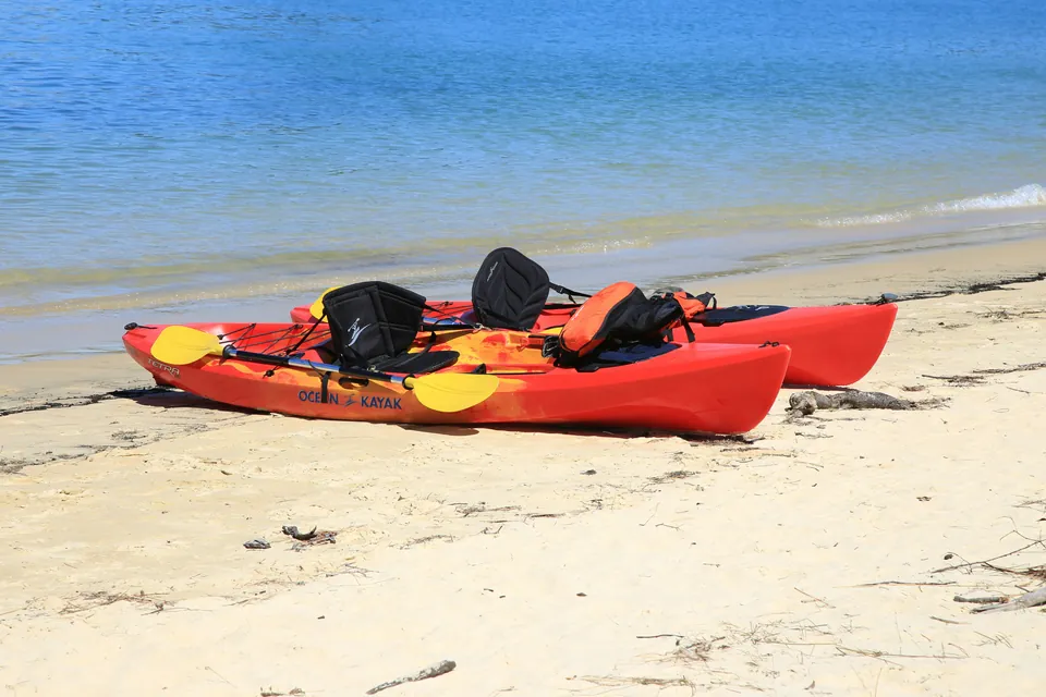 Two red and yellow sit-on-top kayaks with frame seats, paddles and an orange life jacket lie on a white sand beach beside calm blue water.