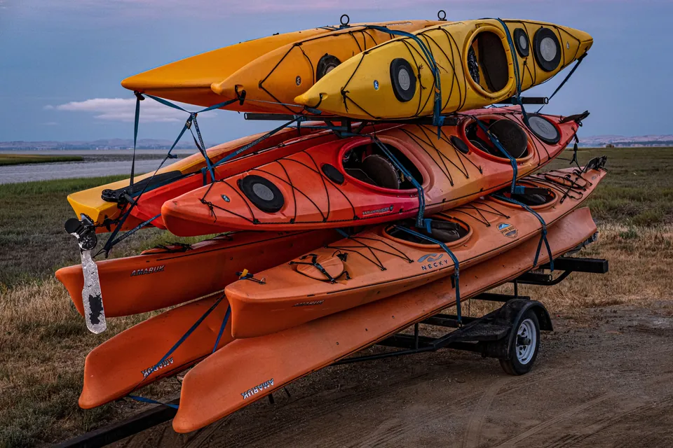 Six orange and yellow kayaks strapped in two tiers on a small trailer beside a flat coastal marsh.