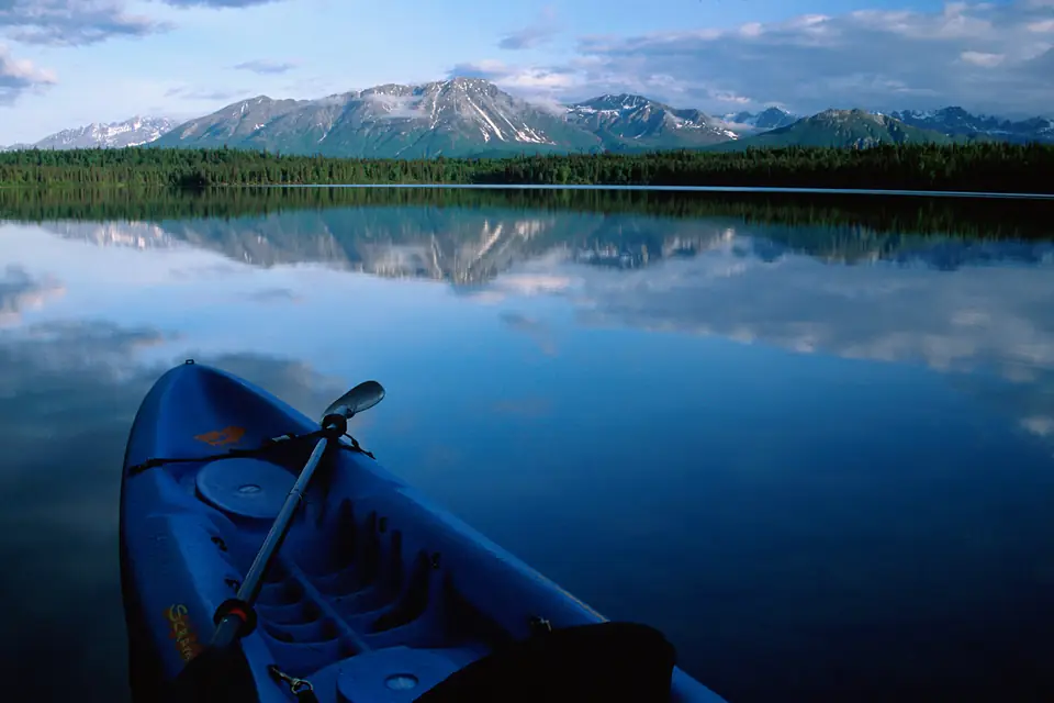 The bow of a blue sit-on-top kayak with a paddle resting across it on a calm lake below snow-capped mountains.