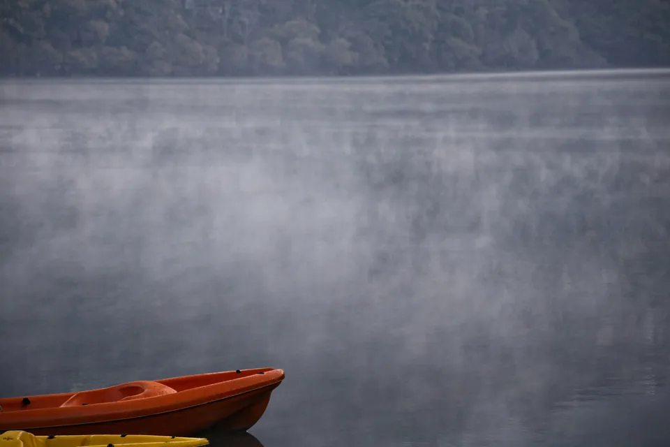 An orange sit-on-top kayak floats at the edge of a still lake with mist rising off the water and a dark forested hillside behind.
