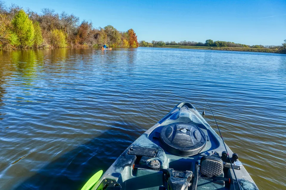 The bow of a rigged fishing kayak on a calm river in autumn, with a bow hatch, gear tracks, a rod holder and a paddle blade in view.