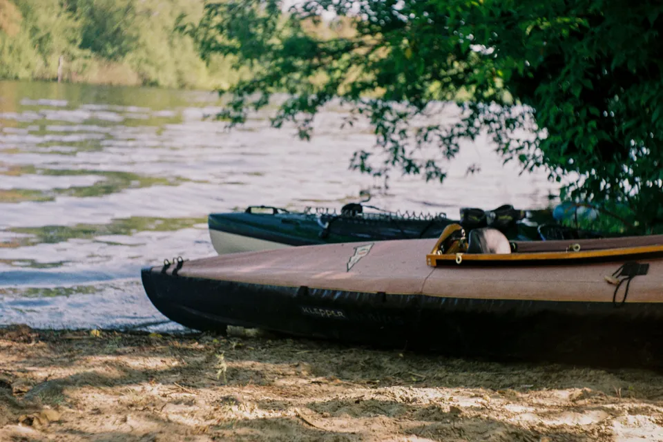 Two kayaks pulled onto a sandy riverbank beneath overhanging trees, with sunlit river water behind them.