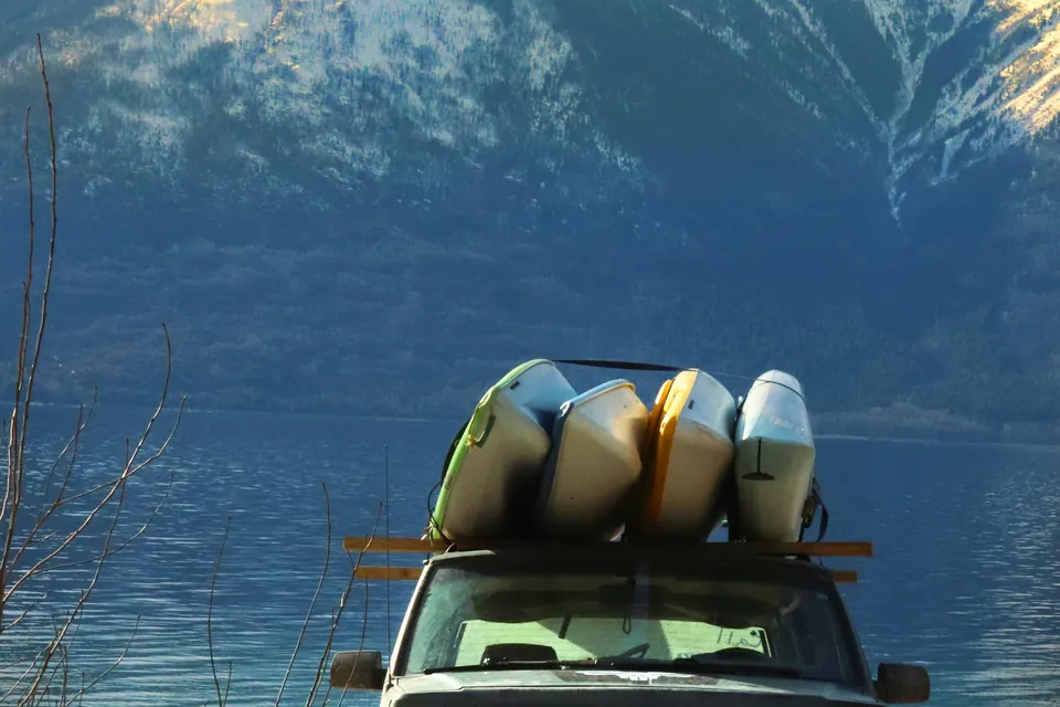 Several kayaks strapped on their sides to a roof rack on a vehicle parked at a lake below snow-capped mountains.