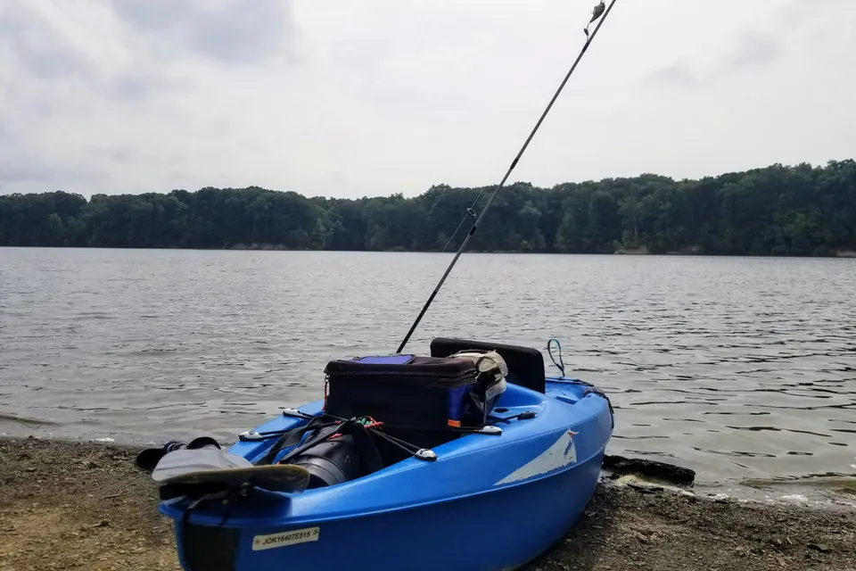 A blue sit-on-top fishing kayak with a tackle bag and a single rod rests on a gravel shoreline, with a calm lake and tree line behind it.