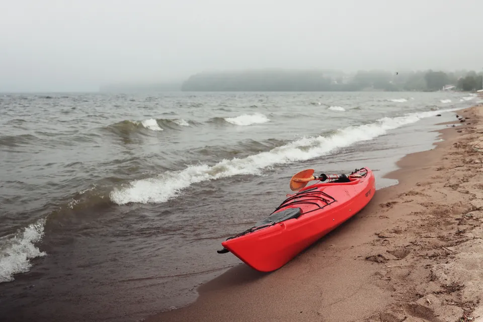 A red kayak pulled up on a sand beach as wind-driven waves break along the shore under a gray sky.