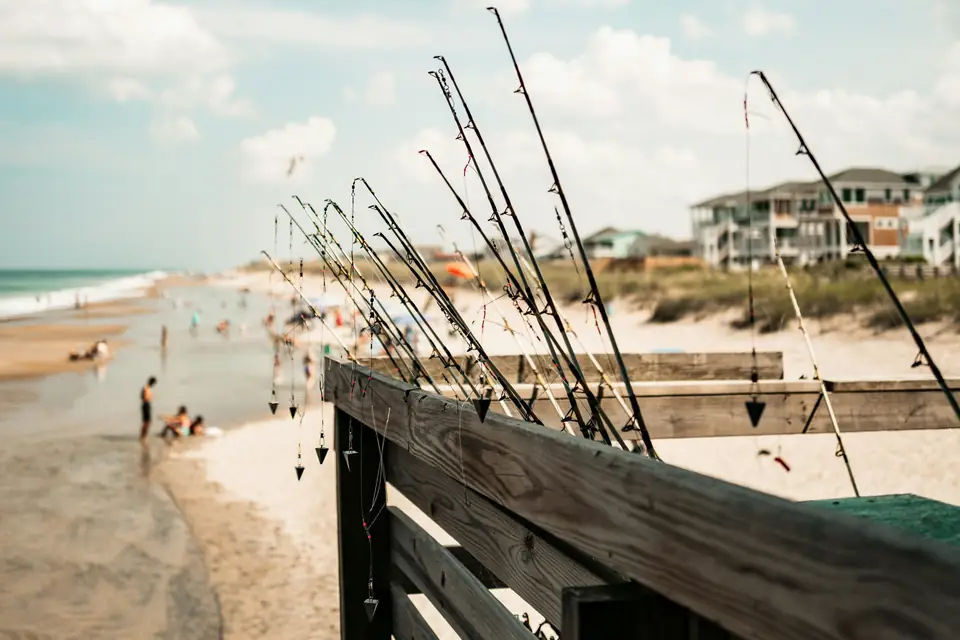 A row of fishing rods leaning on a wooden rail above a beach, each with a pre-tied bottom rig ending in a pyramid sinker hanging from the tip.